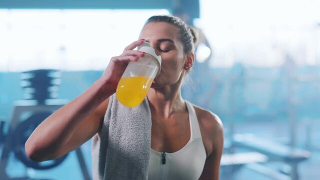 Sexy sweaty young caucasian woman drinking juice from sports cup looking at camera. Fitness girl. Female athlete. After workout. Gym concept.