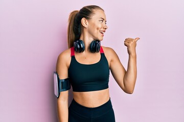 Beautiful young blonde woman wearing gym clothes and using headphones pointing thumb up to the side smiling happy with open mouth