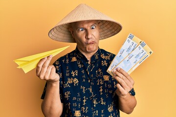Middle age bald man wearing traditional asian hat holding boarding pass making fish face with mouth and squinting eyes, crazy and comical.