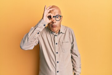 Middle age bald man wearing casual clothes and glasses doing ok gesture shocked with surprised face, eye looking through fingers. unbelieving expression.