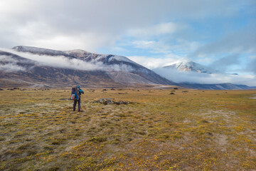 Hiker walking through remote arctic valley on a partly cloudy summer day. Dramatic arctic landscape of Akshayuk Pass, Baffin Island, Canada. Grass and moss in autumn colors.