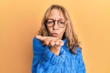 Middle age blonde woman wearing glasses and casual winter sweater looking at the camera blowing a kiss with hand on air being lovely and sexy. love expression.