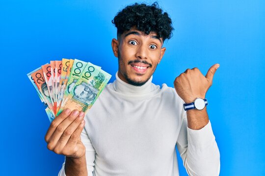 Young arab handsome man holding australian dollars pointing thumb up to the side smiling happy with open mouth