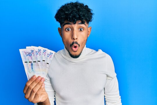 Young arab handsome man holding 50 colombian pesos banknotes scared and amazed with open mouth for surprise, disbelief face