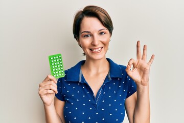 Young brunette woman with short hair holding birth control pills doing ok sign with fingers, smiling friendly gesturing excellent symbol