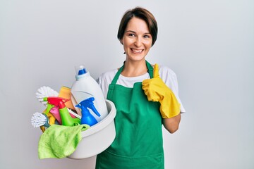 Young brunette woman with short hair wearing apron holding cleaning products doing happy thumbs up gesture with hand. approving expression looking at the camera showing success.