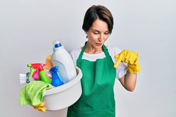Young brunette woman with short hair wearing apron holding cleaning products pointing down with fingers showing advertisement, surprised face and open mouth