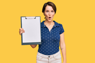 Fototapeta na wymiar Young brunette woman with short hair holding clipboard with blank space scared and amazed with open mouth for surprise, disbelief face