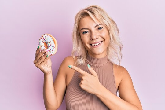 Young blonde girl holding donut smiling happy pointing with hand and finger