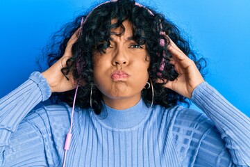 Young hispanic woman listening to music using headphones puffing cheeks with funny face. mouth inflated with air, catching air.