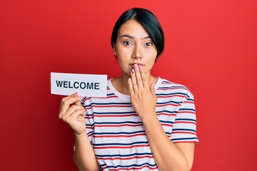 Beautiful young woman with short hair holding welcome paper covering mouth with hand, shocked and afraid for mistake. surprised expression