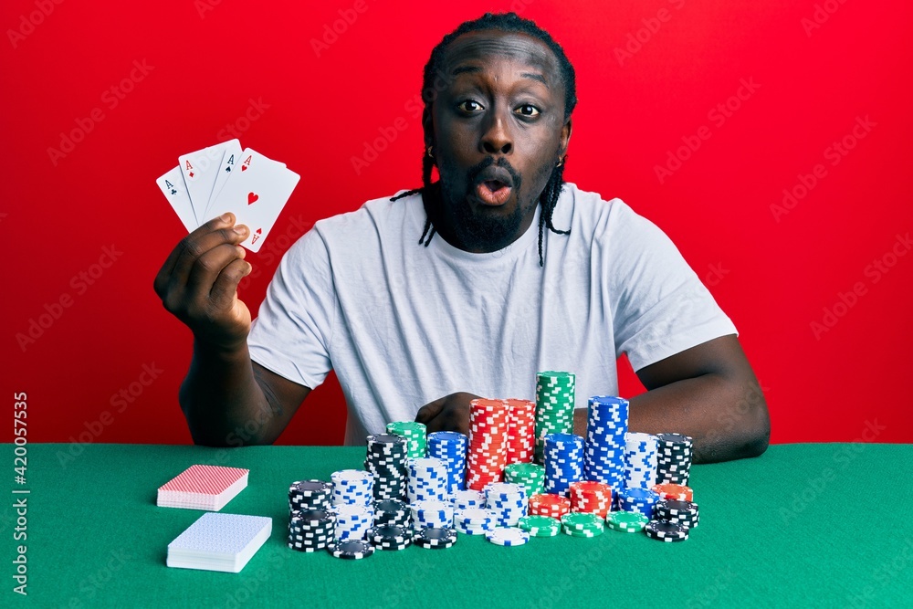 Poster handsome young black man playing poker holding cards scared and amazed with open mouth for surprise,