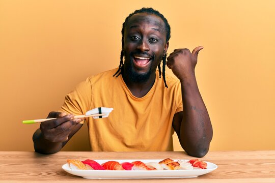 Handsome young black man eating sushi sitting on the table pointing thumb up to the side smiling happy with open mouth