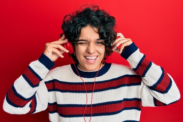 Young hispanic woman with curly hair listening to music using headphones winking looking at the...