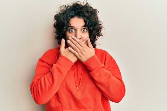 Young hispanic woman with curly hair wearing casual sweatshirt shocked covering mouth with hands for mistake. secret concept.