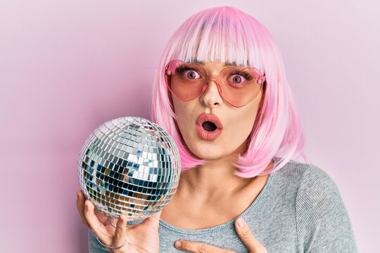 Young caucasian woman wearing pink wig holding disco ball scared and amazed with open mouth for surprise, disbelief face