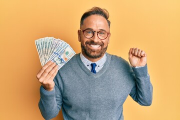 Handsome middle age man holding dollars screaming proud, celebrating victory and success very excited with raised arm