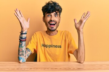 Young hispanic man wearing tshirt with happiness word message sitting on the table celebrating crazy and amazed for success with arms raised and open eyes screaming excited. winner concept