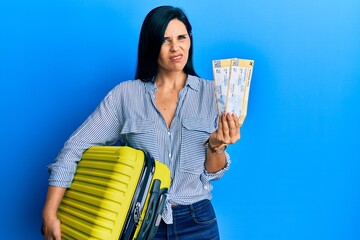 Young caucasian woman holding cabin suitcase and boarding tickets clueless and confused expression....