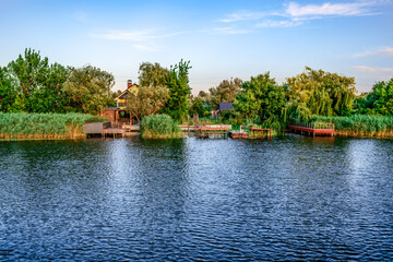 Fototapeta premium Residential houses with personal piers on Konka River coast in Kherson floodplains (Ukraine). Suburban area with buildings on the shore among green trees and reeds on the water surface background