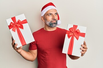 Middle age handsome man wearing christmas hat and holding a gifts smiling looking to the side and staring away thinking.