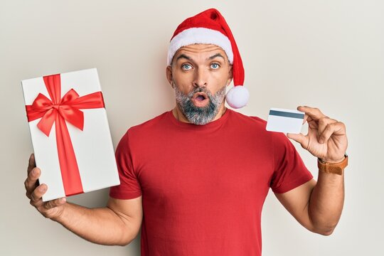 Middle age handsome man wearing chrismast hat holding gift and credit card in shock face, looking skeptical and sarcastic, surprised with open mouth