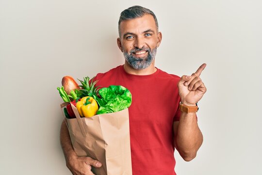 Middle age handsome man holding paper bag with bread and groceries smiling happy pointing with hand and finger to the side