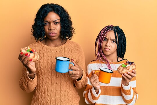 Beautiful african american mother and daughter holding sweet pastries and drinking coffee skeptic and nervous, frowning upset because of problem. negative person.