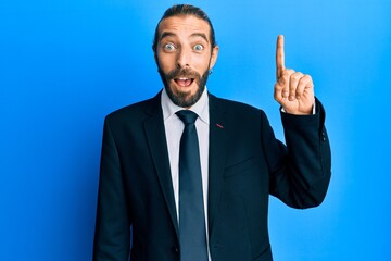 Attractive man with long hair and beard wearing business suit and tie pointing finger up with successful idea. exited and happy. number one.