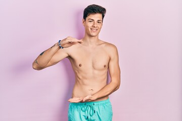 Fototapeta premium Young hispanic man wearing swimwear shirtless gesturing with hands showing big and large size sign, measure symbol. smiling looking at the camera. measuring concept.