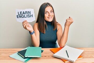 Young caucasian woman holding learn english paper while studying screaming proud, celebrating victory and success very excited with raised arm