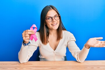 Young caucasian woman holding pink cancer ribbon celebrating achievement with happy smile and winner expression with raised hand