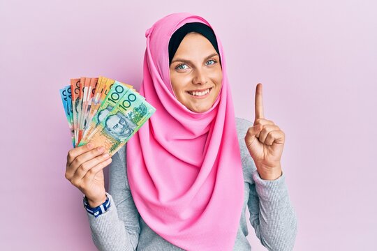 Young caucasian woman wearing islamic hijab holding australian dollars banknotes smiling with an idea or question pointing finger with happy face, number one