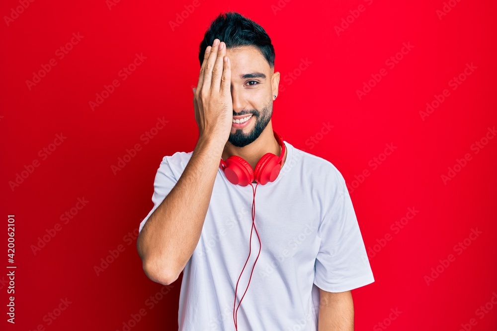 Wall mural young man with beard listening to music using headphones covering one eye with hand, confident smile