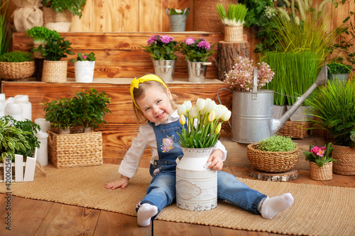 Little girl with a bouquet of white tulips sits on porch of wooden house, around houseplants and flowers. Celebration concept. Gardening. Child playing in backyard in garden. Birthday, mother's day