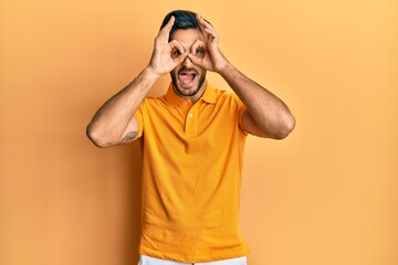 Young hispanic man wearing casual yellow t shirt doing ok gesture like binoculars sticking tongue out, eyes looking through fingers. crazy expression.