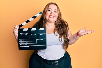 Plus size caucasian young woman holding video film clapboard celebrating achievement with happy...