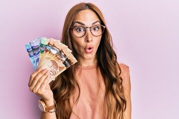 Young brunette woman holding canadian dollars scared and amazed with open mouth for surprise,...