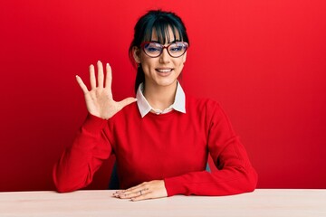 Young brunette woman with bangs wearing glasses sitting on the table showing and pointing up with...