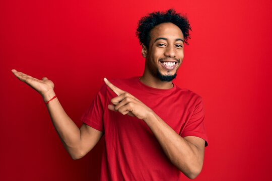 Young african american man with beard wearing casual red t shirt amazed and smiling to the camera while presenting with hand and pointing with finger.