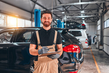 Mechanics at the repair shop. Cheerful young mechanic writing something in his clipboard, cars on the background.