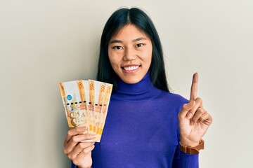 Young chinese woman holding 500 philippine peso banknotes smiling with an idea or question pointing finger with happy face, number one