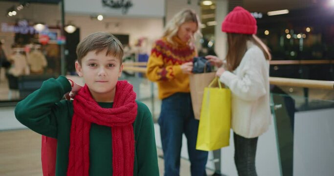 Young smiling teenager boy holding Christmas gift looking posing at camera in shopping mall while his family talking on background. Children concept. Relationships. Holidays.