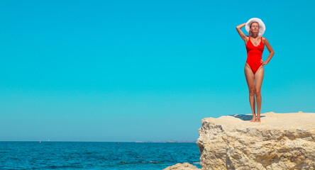 Beautiful young woman in a red swimsuit and white hat posing on white rocks against the backdrop of a bright blue seascape