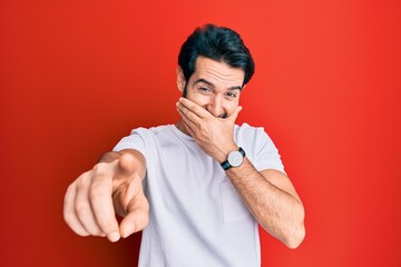 Young hispanic man wearing casual white tshirt laughing at you, pointing finger to the camera with hand over mouth, shame expression