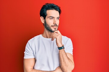 Young hispanic man wearing casual white tshirt with hand on chin thinking about question, pensive expression. smiling and thoughtful face. doubt concept.