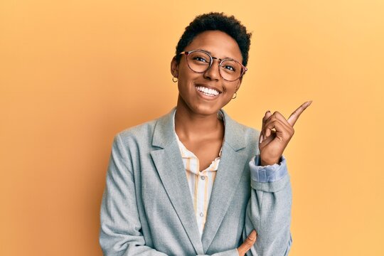 Young african american girl wearing business jacket and glasses with a big smile on face, pointing with hand and finger to the side looking at the camera.