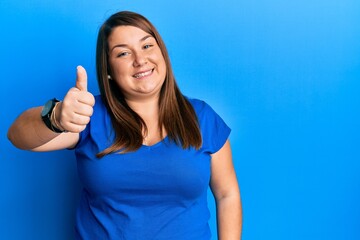 Fototapeta premium Beautiful brunette plus size woman wearing casual blue t shirt doing happy thumbs up gesture with hand. approving expression looking at the camera showing success.