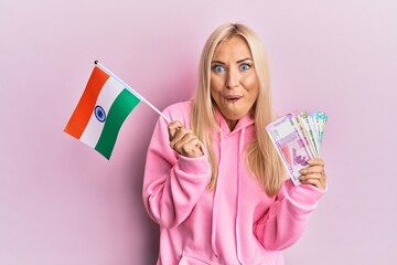 Young blonde woman holding india flag and rupee banknotes afraid and shocked with surprise and amazed expression, fear and excited face.