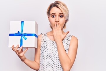 Young beautiful blonde woman holding gift covering mouth with hand, shocked and afraid for mistake....
